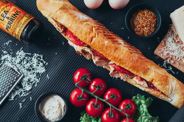 Long baguette sandwich with lettuce, vegetables, salami, chili and cheese on black background