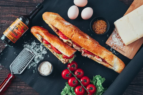 Long baguette sandwich with lettuce, vegetables, salami, chili and cheese on black background
