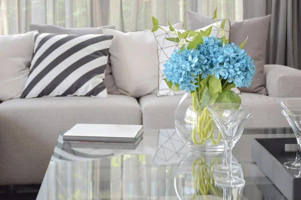 Light blue flower and wine glasses on center table with striped black and white pillow and gray tone pillows on beige sofa