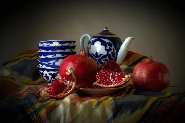 Oriental motifs. Tea pot, bowls, whole and cut pomegranate fruit on a plate. all this on the table, which is covered with traditional Asian cloth.
