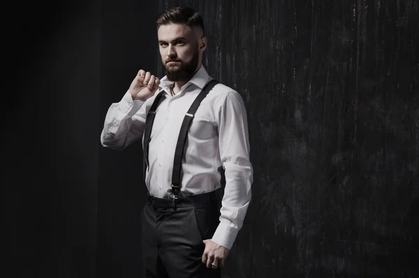 Attractive bearded man in a white shirt and suspenders standing near dark wall.