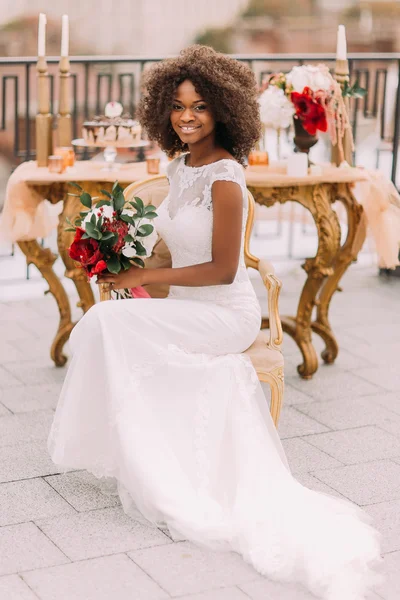 Happy young black bride smiling with bouquet of red flowers and sitting on vintage terracotta chair