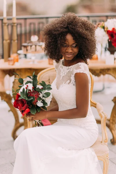 Wonderful black bride happily smiling with eyes closed and holding a bouquet of red flowers. Wedding day