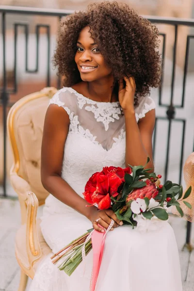 Happy young black bride smiling with bouquet of red flowers and sitting on vintage terracotta chair