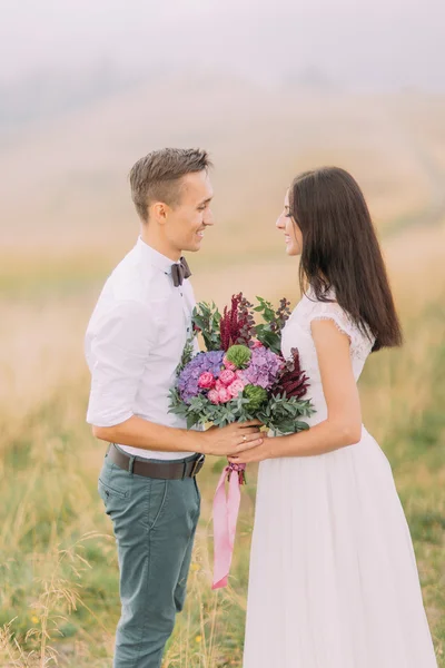 Happy bride and groom with bouquet in hands lovingly look at each other. Misty field on background