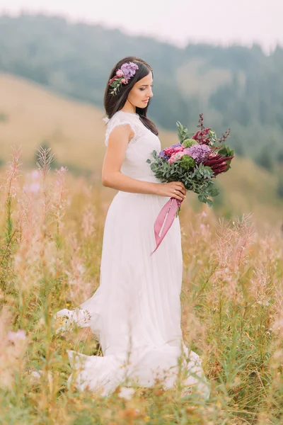 Lovely brunette bride standing with bouquet on the field in Alpian mountains
