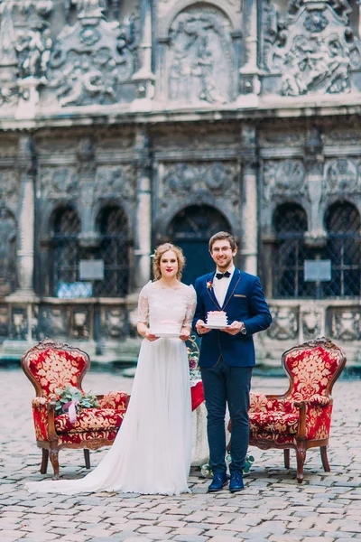 Charming newlyweds holding pieces of their wedding cake and smiling. Old european architecture on background.