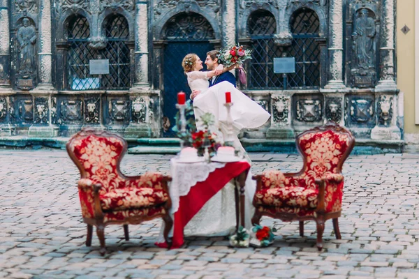 Cheerfull groom in glasses  holds in hands his blonde bride  near the romantic table for two setted for them.