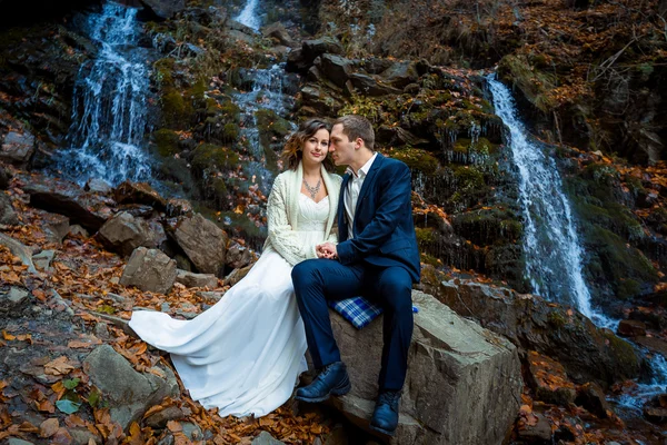 Wedding couple sitting on the stone and posing. Waterfall in mountains background. Fall