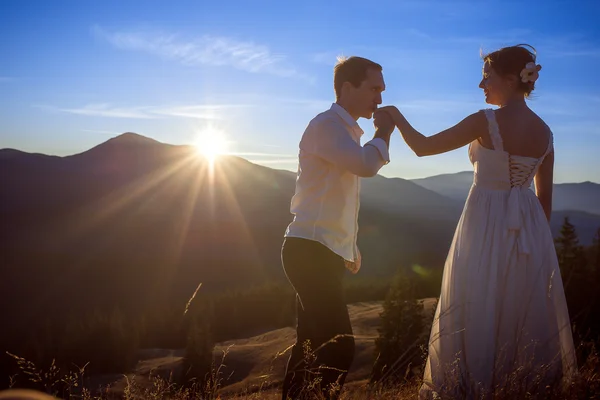 Groom kisses hand of bride. Sunset in the mountains on background.