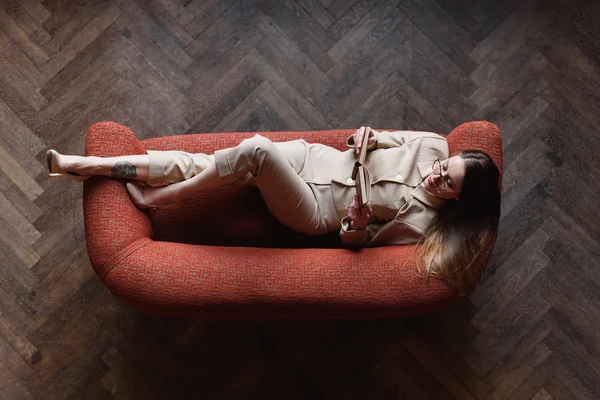 Stylish young girl in beige suit reads a book on the red sofa. Top view