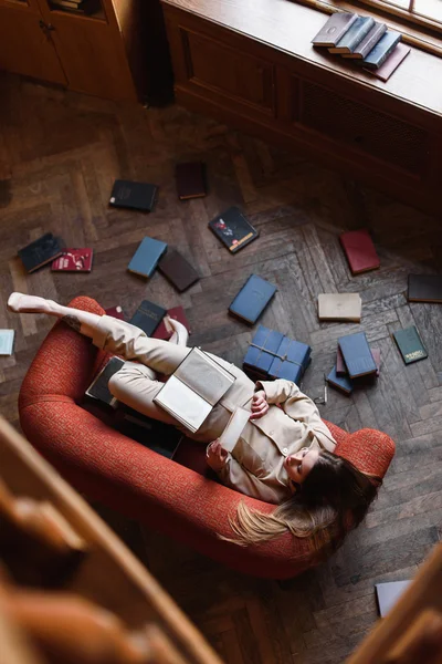 Stylish young girl in beige suit reads a book on the red sofa surrounded by books. Top view