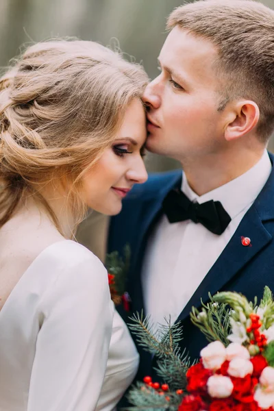 Close up portrait of stylish groom kissing his beautiful bride in forehead. Sunny day at the forest