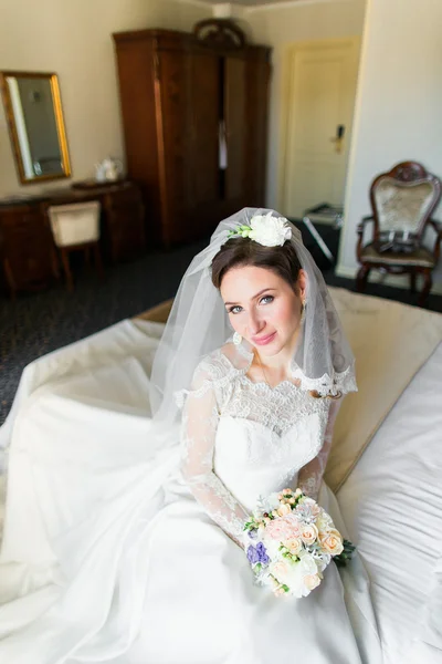Beautiful young bride with makeup, fancy hairstyle in white dress and veil sitting on bed