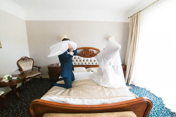 Portrait of happy smiling newlywed couple fighting with pillows on bed in hotel room