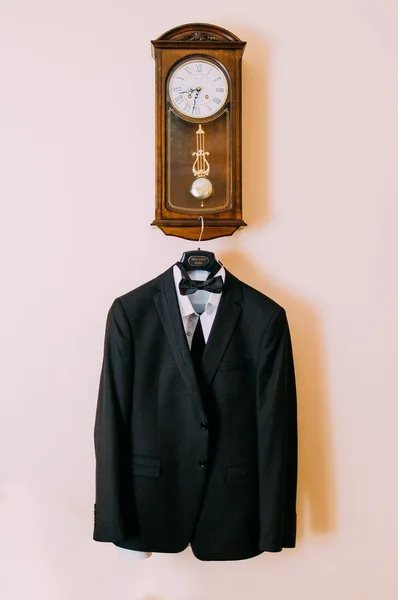 Elegant stylish black wedding suit hanging on wooden clock hotel room