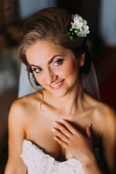 Smiling blonde bride in veil and pearl necklace touching her neck indoors