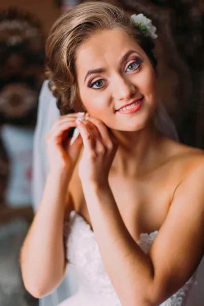 Close-up portrait of beautiful bride in white wedding dress puting on earring and looking at the camera
