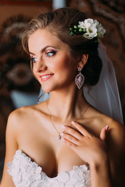 Smiling blonde bride in veil and pearl necklace touching her neck indoors