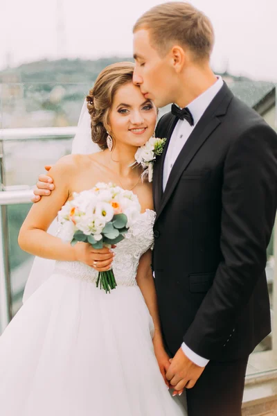 Happy beautiful couple, groom kissing bride forehead on the terrace with cityscape background