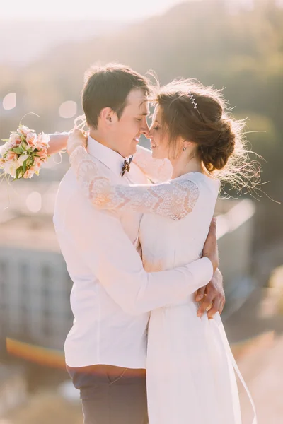 Newlyweds touch each other with noses, while bride holds  bouquet on cityscape background