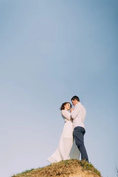 Happy beautiful bride and groom embracing face-to-face at top of hill against to the sky