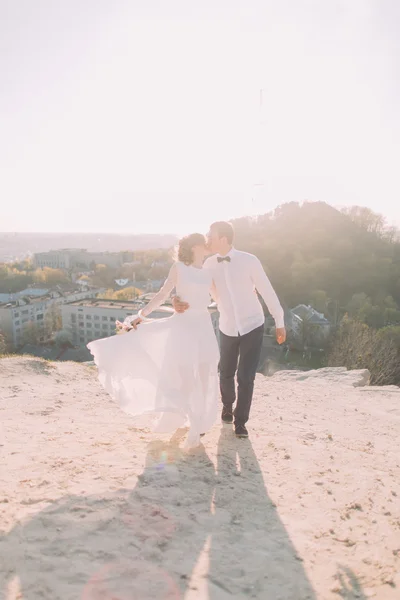 Handsome groom in white shirt and bride wearing bridal dress walking kissing against the backdrop of city