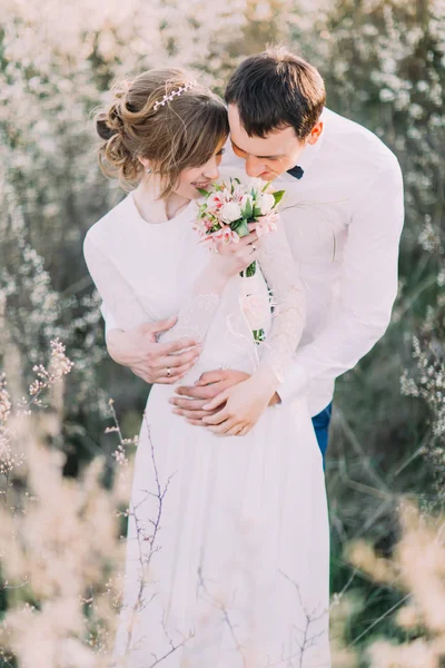 Handsome groom gently holds the bride back holding bouquet in the blossoming spring garden