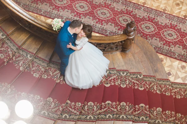 Handsome elegant groom kissing cheek of beautiful smiling bride on old wooden stairs at the rich interior  background