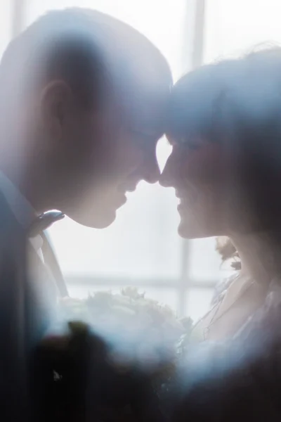 Silhouette of bride and groom embracing holding bouquet against the window with curtains, close-up