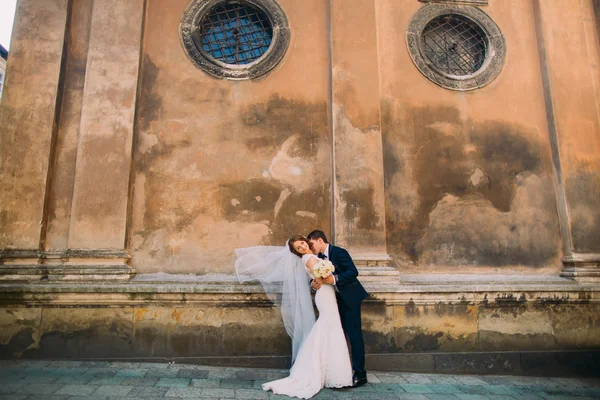 Groom kisses brides neck tenderly holding her waist near brown wall of old church