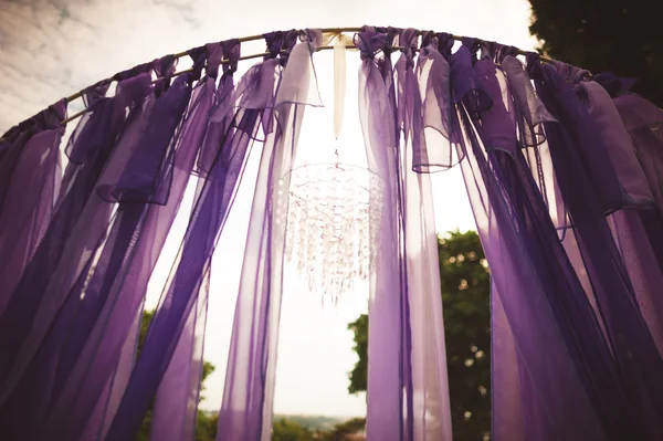 Wedding arch decorated with purple cloth ribbons waving in the wind and luxury chandelier on background