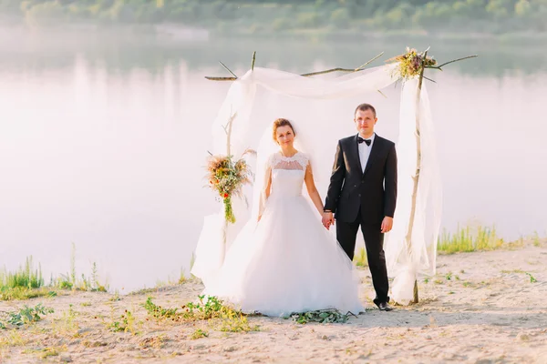 Beautiful bride in white dress and handsome groom wearing black suit standing under archway on beach wedding ceremony near lake