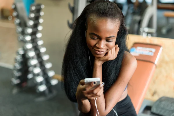 Portrait of gorgeous black lady with luxury long hair texting on her smartphone in the gym