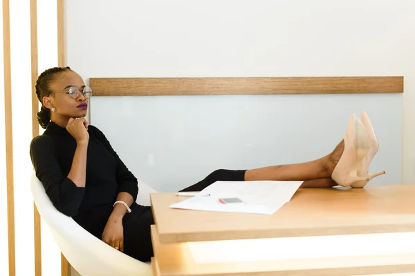 Elegant African or black American business woman, wearing glasses, sitting with legs in beige stilettos pumps shoes on table and thinking