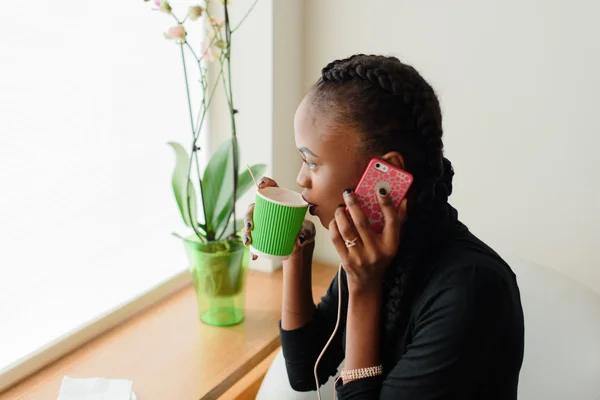 Smart black american woman talking on phone holding disposable cup next to window