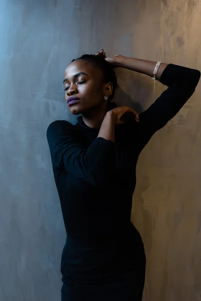 Close up portrait of beautiful young woman in black dress with eyes closed and hands over head, gray background