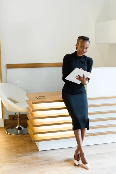 Stern elegant business woman wearing black dress and beige shoes in light office looking at her agenda, full length portrait