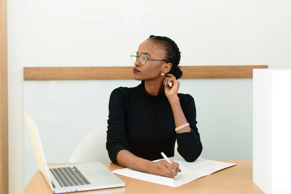 Thoughtful black businesswoman in glasses touching her neck looking away at desk with notepad and laptop in office