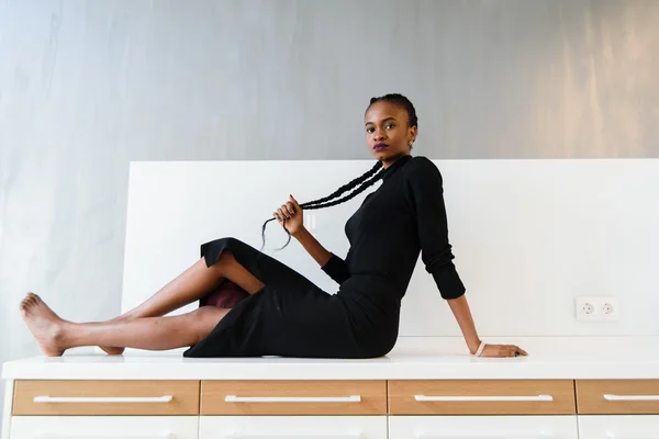 Elegant african or black american woman in dark dress posing on desk in light interior