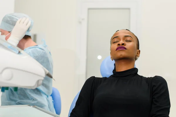 African-American ethnic black patient sitting closing eyes waiting, while her dentist preparing to operation on background
