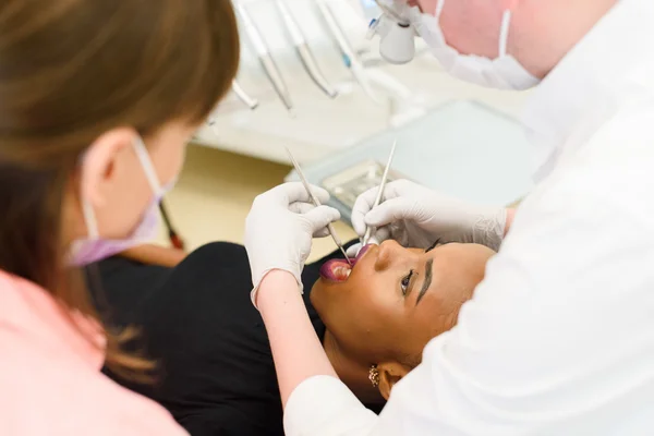 Young African-American ethnic black female opening mouth while dentist in white latex gloves and mask, his assistant check condition of her teeth