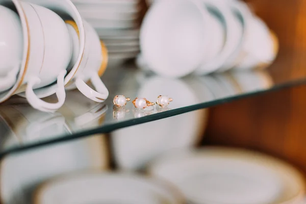 Extreme close-up gold jewelry embellished with pearls on glass cupboard shelf. Porcellain cups and plates at background