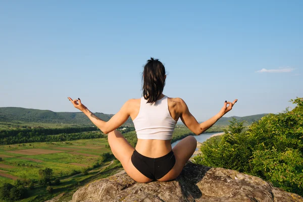 Back view of yoga woman with perfect muscular body meditating in lotus posture on the rock. Mountain landscape and blue sky background