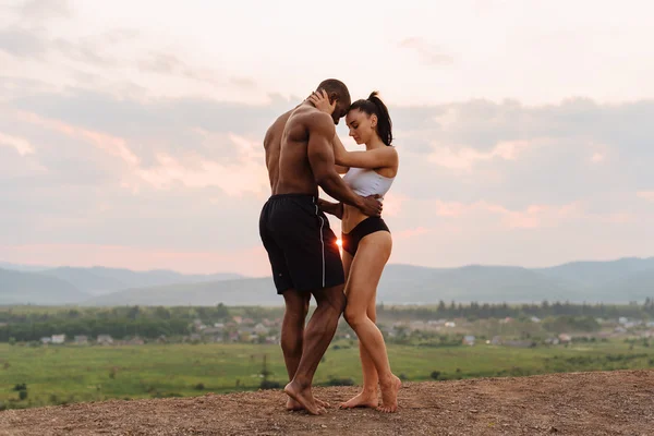 Full-length fashion portrait of sexy fit mixed race couple with perfect bodies in sportswear softly embracing on mountains landscape background. Beauty of humans body