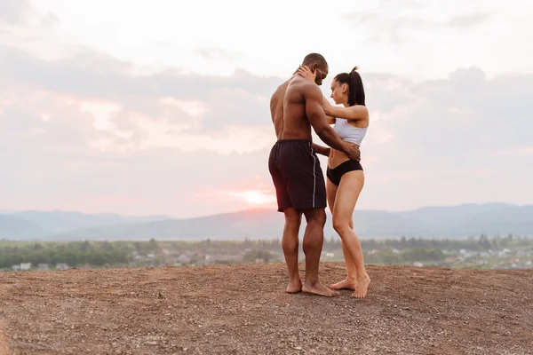 Full-length portrait of sexy fit mixed race couple with perfect bodies in sportswear softly embracing on mountains landscape background.