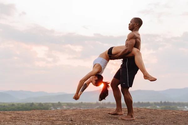 Mixed race gymnastic couple with perfect bodies in sportswear dancing on mountains landscape background. Pink sunset sky and white clouds