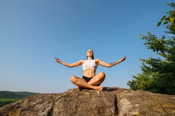 Sexy young athletic woman with muscular body practicing yoga on the rock. Blue sky background. Moment of harmony and relaxing