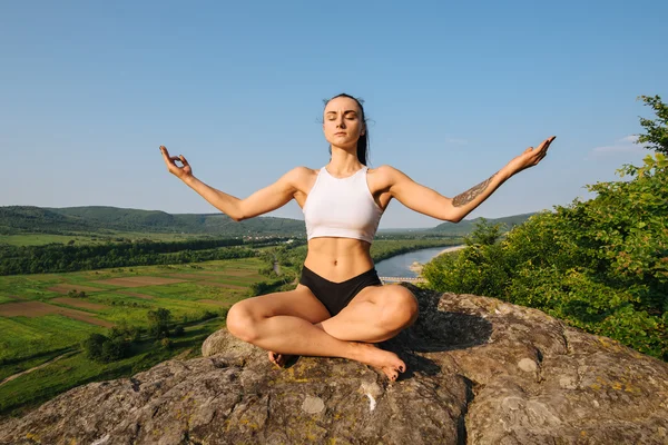 Sexy young brunette woman with muscular body practicing yoga on the rock. Blue sky background. Harmony and relaxing concept