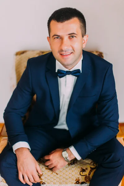 Stylish and confident young handsome man in dark blue suit with bow-tie sitting in vintage chair against grey background